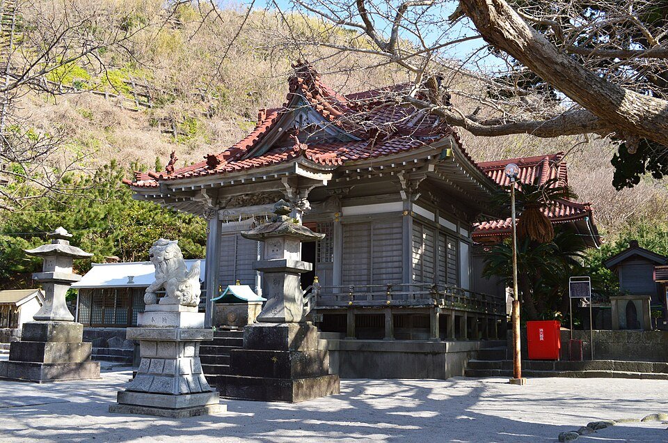赤い瓦屋根の物忌奈命神社の社殿と、手前に立つ狛犬・石灯籠、白い砂が広がる境内