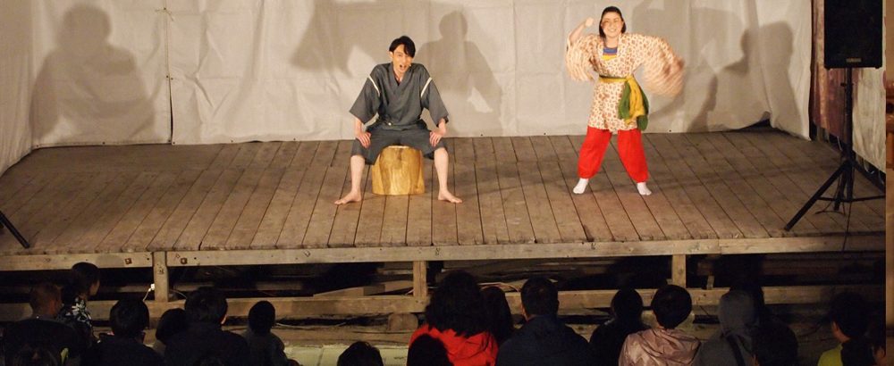 夜の野外特設舞台で作務衣姿の男性と和装の女性が語る「にっぽん御岳話」の公演