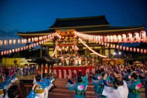 西新井大師の本堂前に組まれた櫓を提灯が囲み、青い浴衣の踊り手が輪になって手を挙げている夜の様子