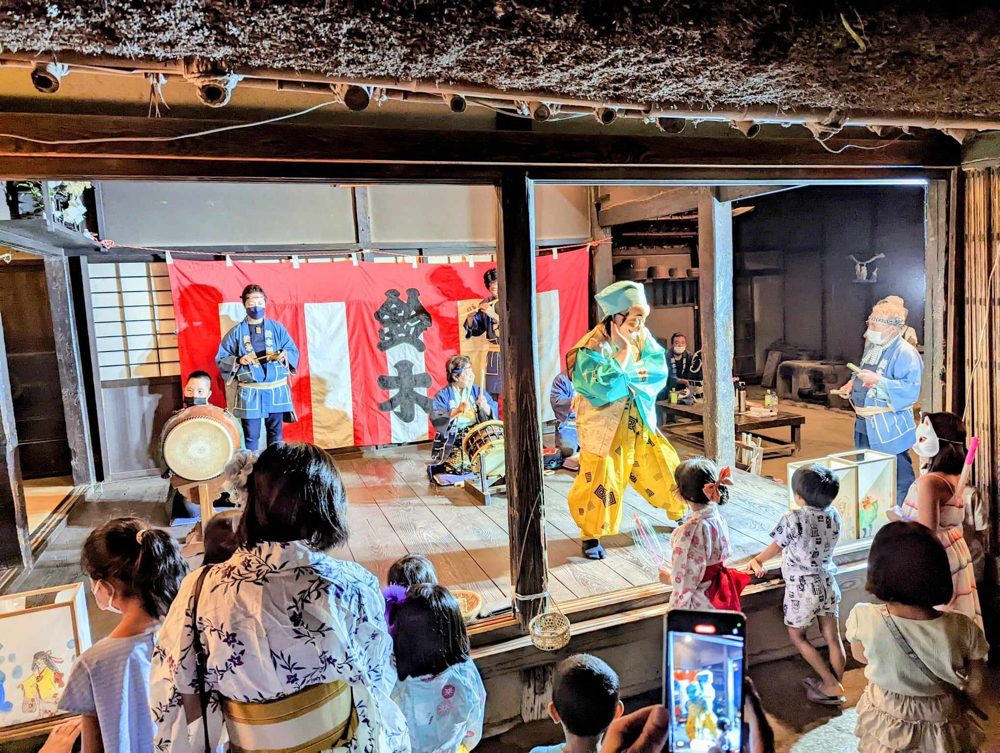 茅葺き古民家の板の間で行われる鈴木ばやしの実演と、浴衣姿で見物する子どもたち