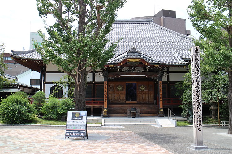 緑に囲まれた天妙国寺の本堂正面。瓦屋根、木造の扉、境内入口の石柱が写っている
