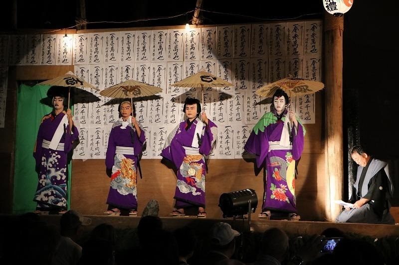 夜の野外特設舞台で、紫の着物に白塗りの演者4人が番傘を持って並ぶ。背景の壁は「金壱萬圓也」などと墨書された奉納札で埋め尽くされ、右手に黒紋付の人物が台本を持って立つ