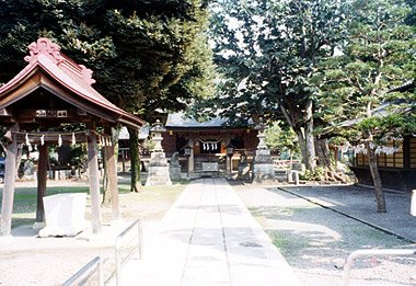 諏訪神社の境内。左手前に赤い屋根の手水舎、中央に石畳の参道がまっすぐ奥へ伸び、突き当たりに社殿。右手に大きな常緑樹が枝を広げている。人の姿はない