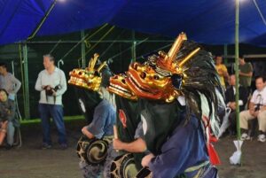 夜、青いテントの下で電球に照らされ、金色の角と赤い顔を持つ獅子頭に黒い長毛をつけた舞い手が3人並んで舞う。腹に締太鼓を抱えている。周囲にパイプ椅子で見物する人たち