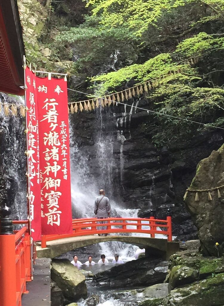 犬鳴山七宝瀧寺の行者ヶ瀧。赤い欄干の橋の下の滝壺に白衣姿の4人が浸かり、橋の上に僧侶が立つ