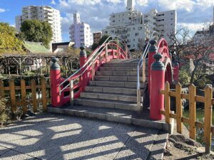 亀戸天神社の心字池に架かる朱塗りの太鼓橋。石段と朱の擬宝珠、左手に藤棚、背景にマンション群と青空