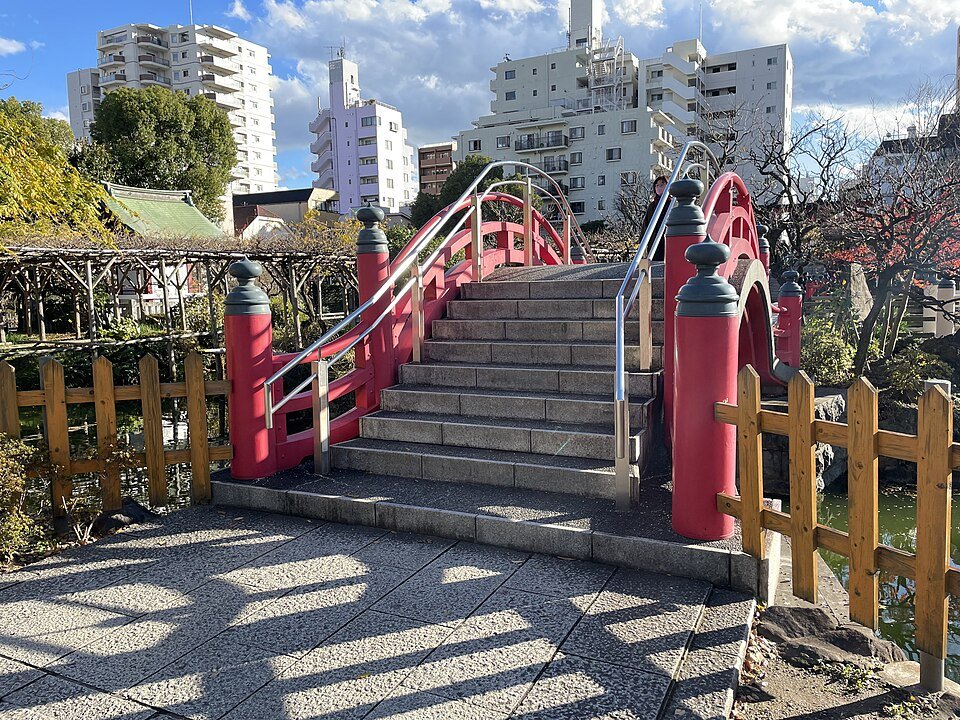 亀戸天神社の心字池に架かる朱塗りの太鼓橋。石段と朱の擬宝珠、左手に藤棚、背景にマンション群と青空