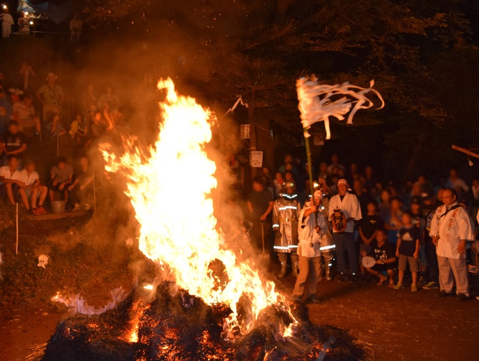 夜の富士塚の麓で円錐形に積まれた麦わらの大松明が高く炎を上げ、斜面に腰かけた見物客が見上げている