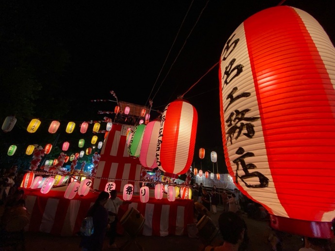 「火の花祭り」と書かれた提灯を下げた紅白幕の櫓と、夜空に張り渡された色とりどりの提灯、櫓の上の太鼓