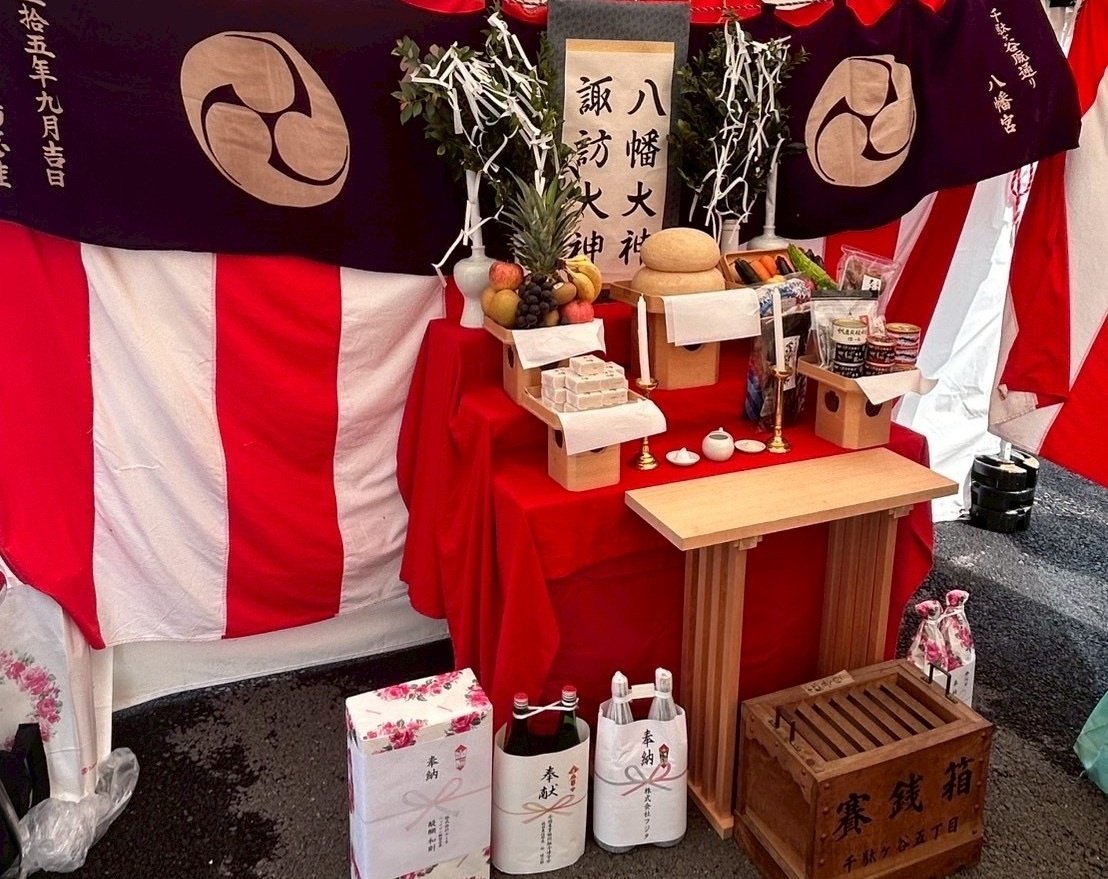千駄ヶ谷五丁目の神酒所。八幡大神・諏訪大神の掛軸の前に鏡餅と果物・野菜が供えられ、奉納の一升瓶と木の賽銭箱が並ぶ