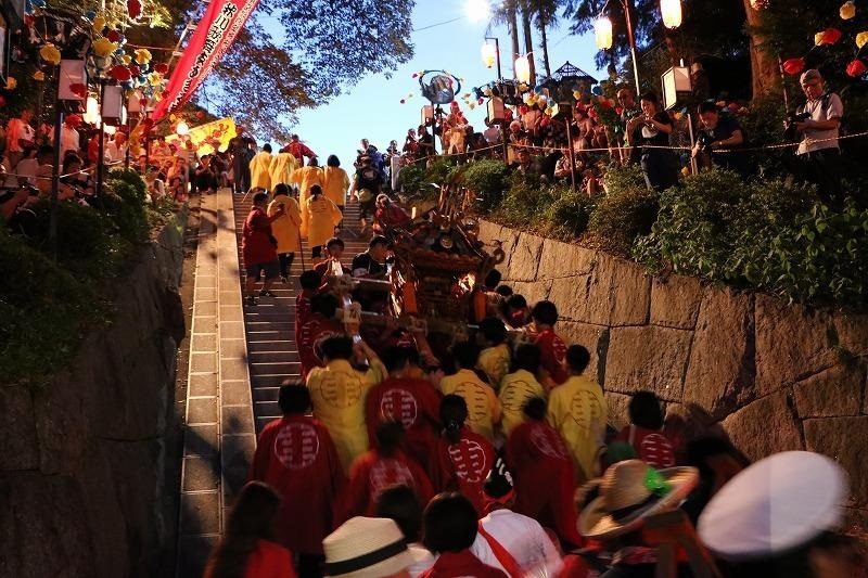 夕暮れ、参道の石段を赤と黄色の法被を着た担ぎ手が埋め尽くし、神輿を担いで上がっていく。両脇の斜面には提灯が連なり、見物客が見下ろしている