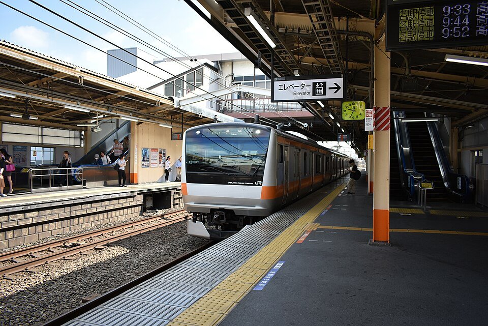JR中央線・西八王子駅のホームに停車するオレンジ帯の電車と、エスカレーター、発車標