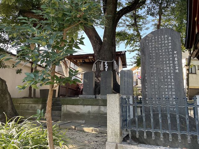 境内に立つ黒御影石の碑。鉄柵に囲われ、縦書きで「女塚古墳の由緒」と刻まれ、碑文に「南北朝時代の烈士新田義貞の子義興公」「侍女少将局」「正平十三年」「矢口の渡し」の文字が読める。奥に注連縄を巻いた大きな御神木と、御即位記念などと刻まれた小さな石碑が3基並ぶ
