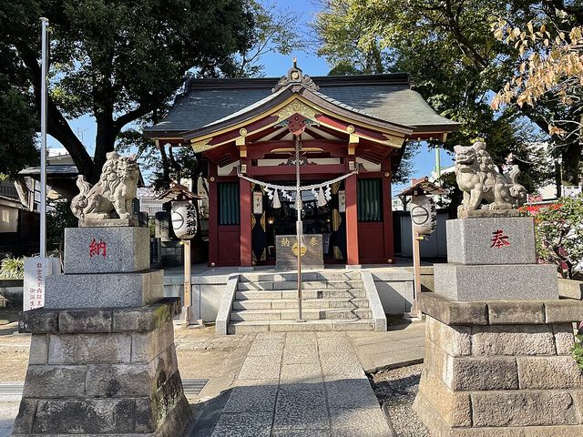 神社の参道から社殿を望む。手前に「納」「奉」と朱字で刻まれた御影石の門柱2基があり、その上に岡崎型の狛犬が一対。奥に朱塗りの社殿、左右に白い提灯。石畳の参道と数段の階段。左手に大きな樹木、右手に低木の植栽と青空