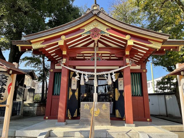 朱塗りの社殿を正面から見た写真。唐破風の彫刻は金・緑・朱の極彩色で、中央に太い注連縄と白い鈴緒が下がる。扉は黒地に金の飾り金具、左右に「奉納 令和」と書かれた白い提灯が2つ。手前に「納」「奉」と記した木製の賽銭箱、下に御影石の石段。左手前に「女塚神社」の赤提灯、背景に大きなケヤキと青空