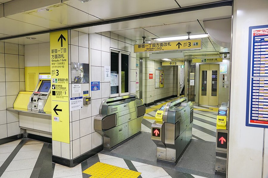 地下鉄駅の改札。黄色い案内サインに「新宿一丁目方面改札 Shinjuku 1 Gate 出口3」と書かれ、自動改札機が3通路並ぶ。左に券売機、右にエレベーター、床は黄と黒のタイル