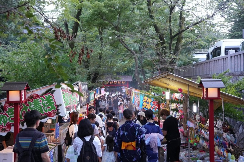 夕方の菅原神社の参道。両側に露店がびっしり並び、浴衣姿の親子連れや学生で通路が埋まっている