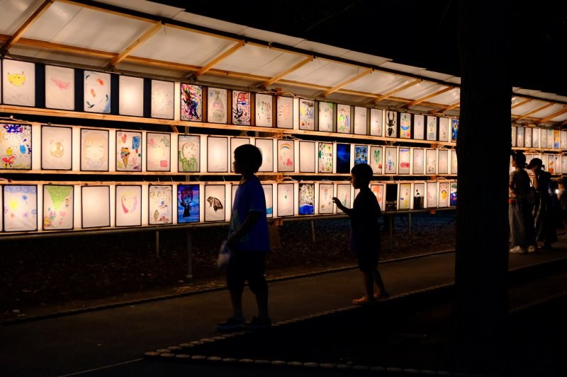夜の菅原神社。屋根付きの木製ラックに子どもの描いた絵灯篭が三段でびっしり並び、内側から光っている。子ども二人が見上げている
