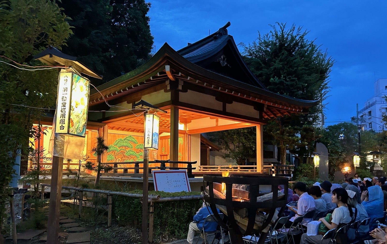 薄暮の鳩森八幡神社能楽殿。舞台手前に篝火の鉄籠が据えられ、後座の壁に老松が描かれ、観客がパイプ椅子に座って見ている