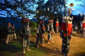 薄暮の土の広場で、青海波柄の藍色の浴衣を着た3人が腹に締太鼓を吊るし、赤と金の毛を垂らした獅子頭をかぶって並んで立つ。手前右に赤白の筒状の被り物をかぶったささら役、左に笛を吹く2人。頭上には紙垂を下げた注連縄が張られ、背景に重なる山の稜線と暮れかけた青空が見える